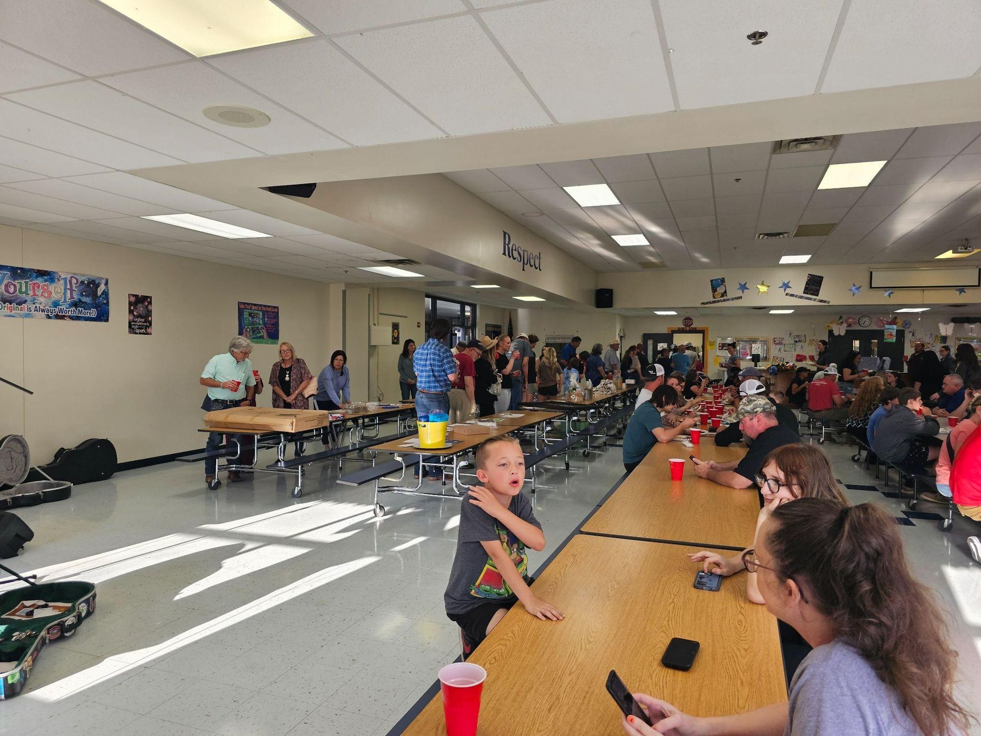 Crowded cafeteria with people seated at long tables and a serving line in the background