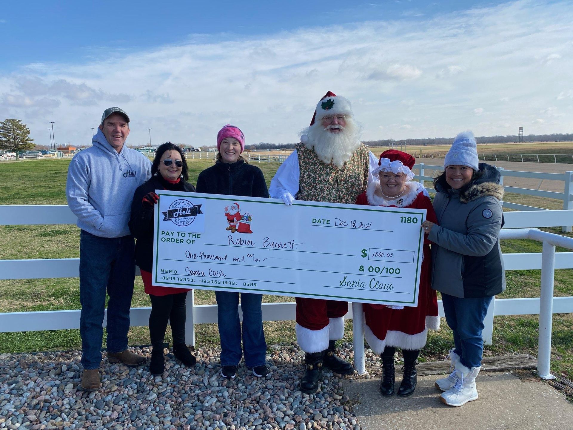 Five people holding an oversized check outdoors, with a Santa figure and a snowman in the background.