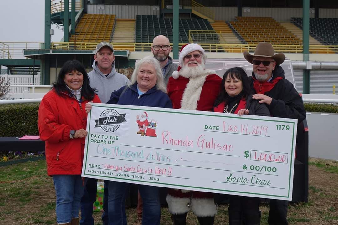 Group holding a large check in a stadium, with Santa costumes and red jackets, smiling at the camera