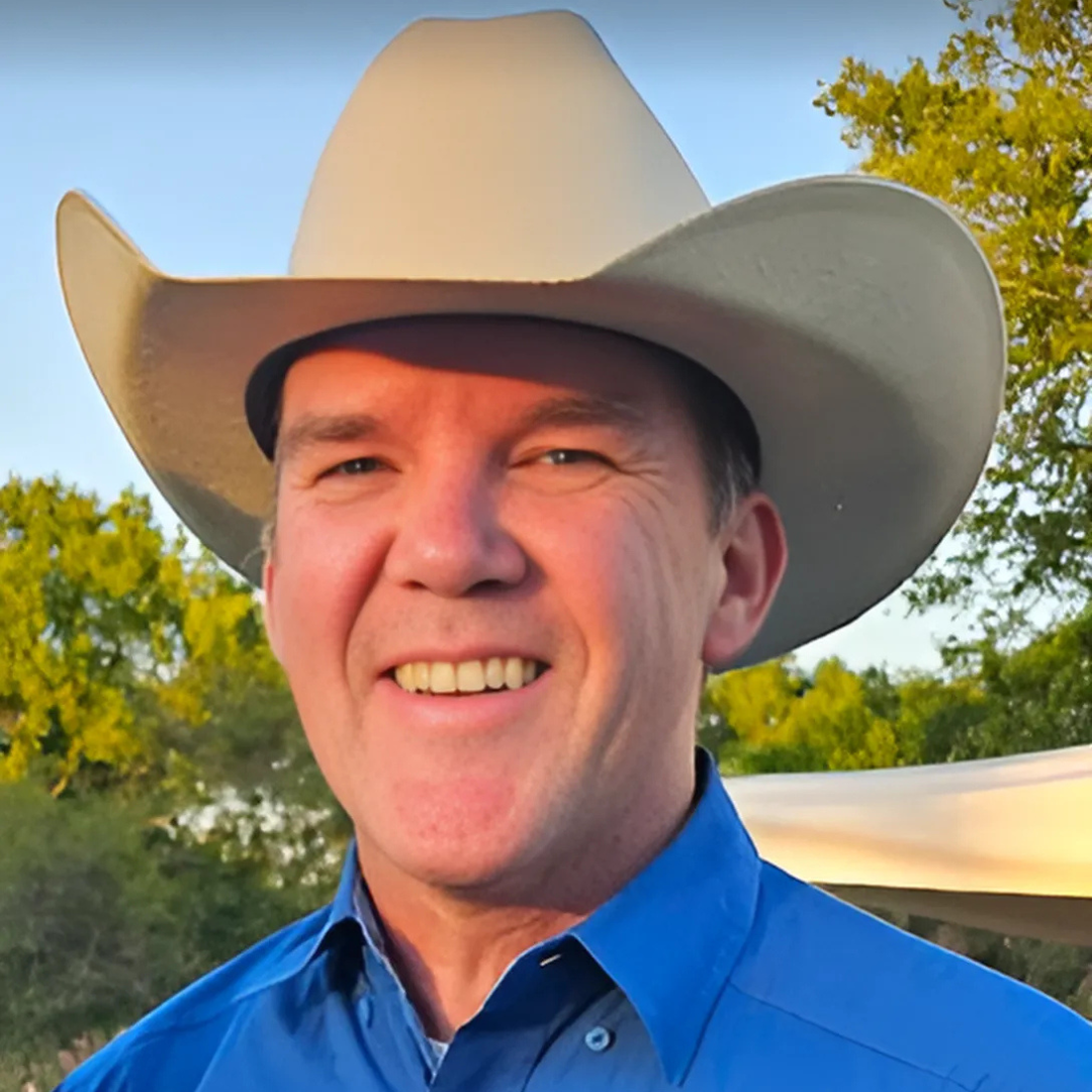 Smiling man in a white cowboy hat and blue shirt outdoors at sunset