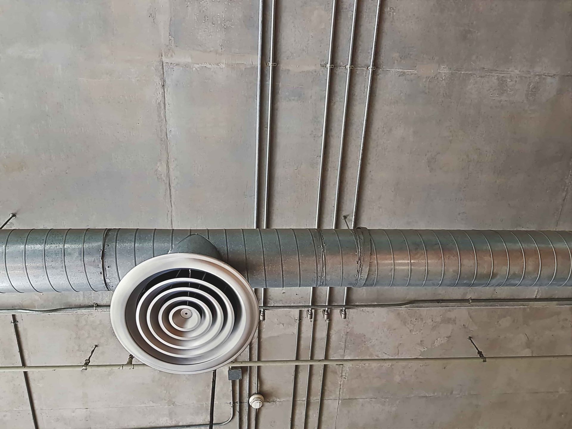 Ceiling duct with a round air vent against a concrete ceiling with exposed pipes