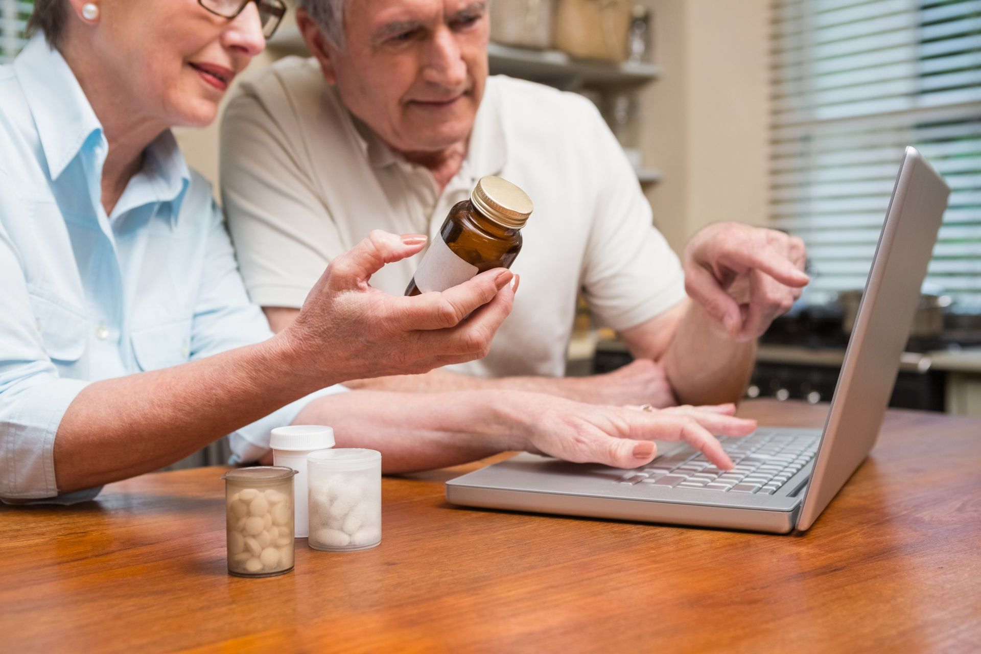 Two people comparing medicine bottles while using a laptop at a wooden table