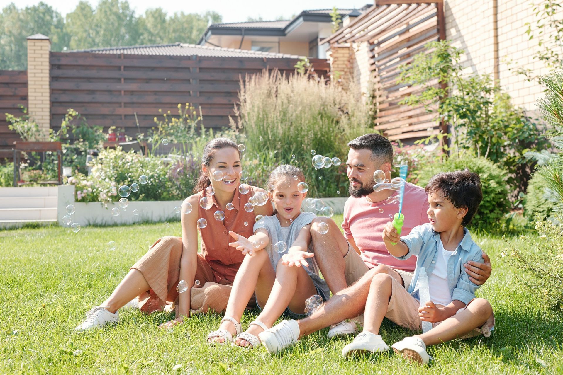 Family sitting on grass in a sunny backyard, smiling and talking together.