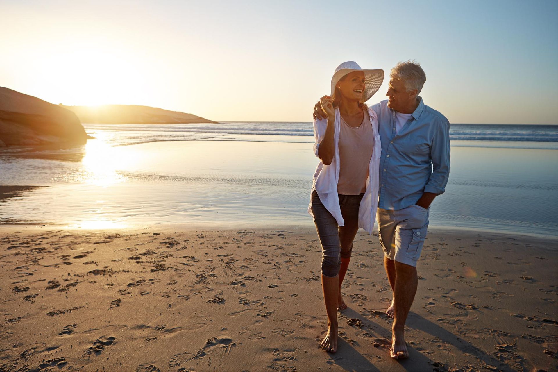 Two people walking barefoot on a beach at sunset, smiling and embracing.