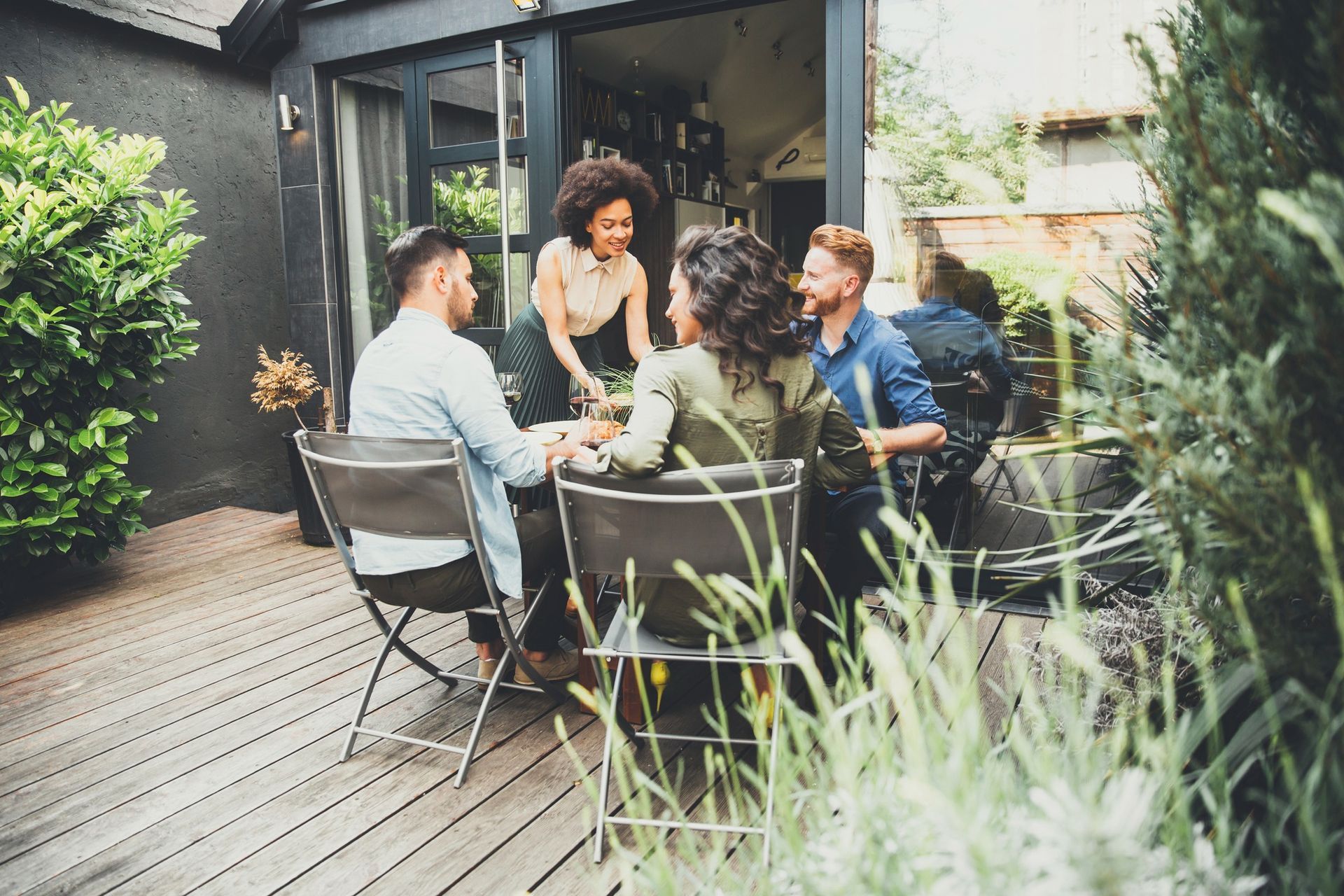 Friends chatting around a table on a patio, with one person serving drinks in a garden setting.