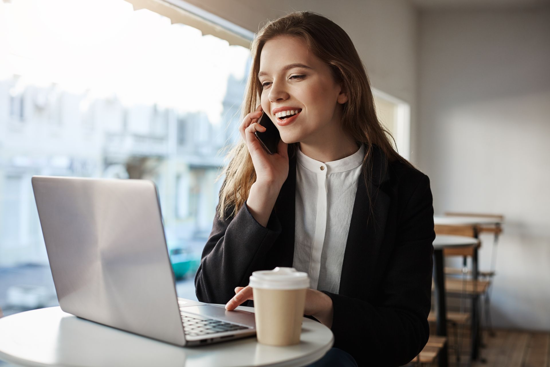 Woman smiling on a laptop in a café, holding a phone beside a takeaway coffee cup.