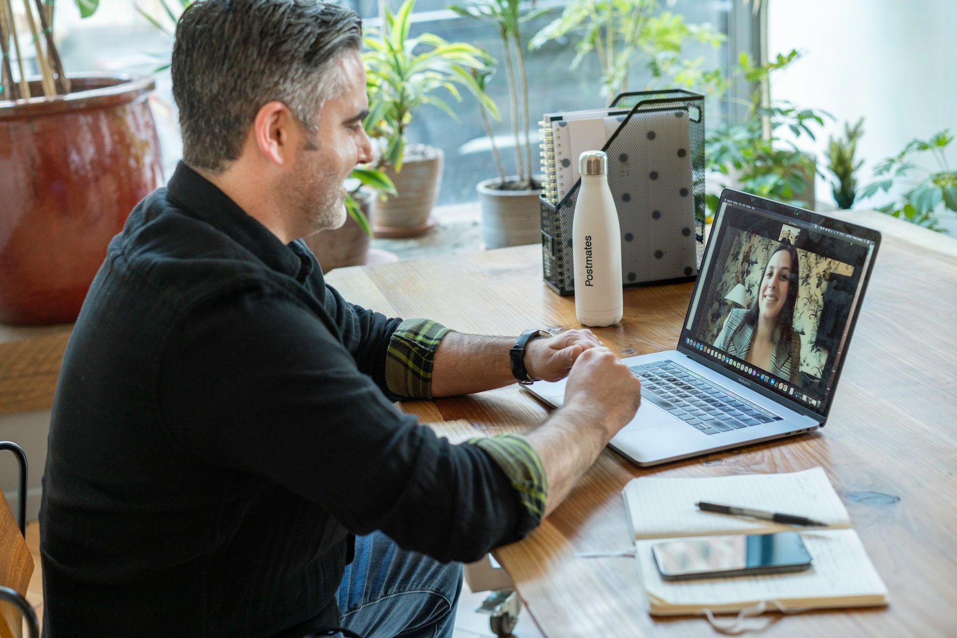 Man using a laptop at a wooden desk in a bright home office, looking at a video call on screen