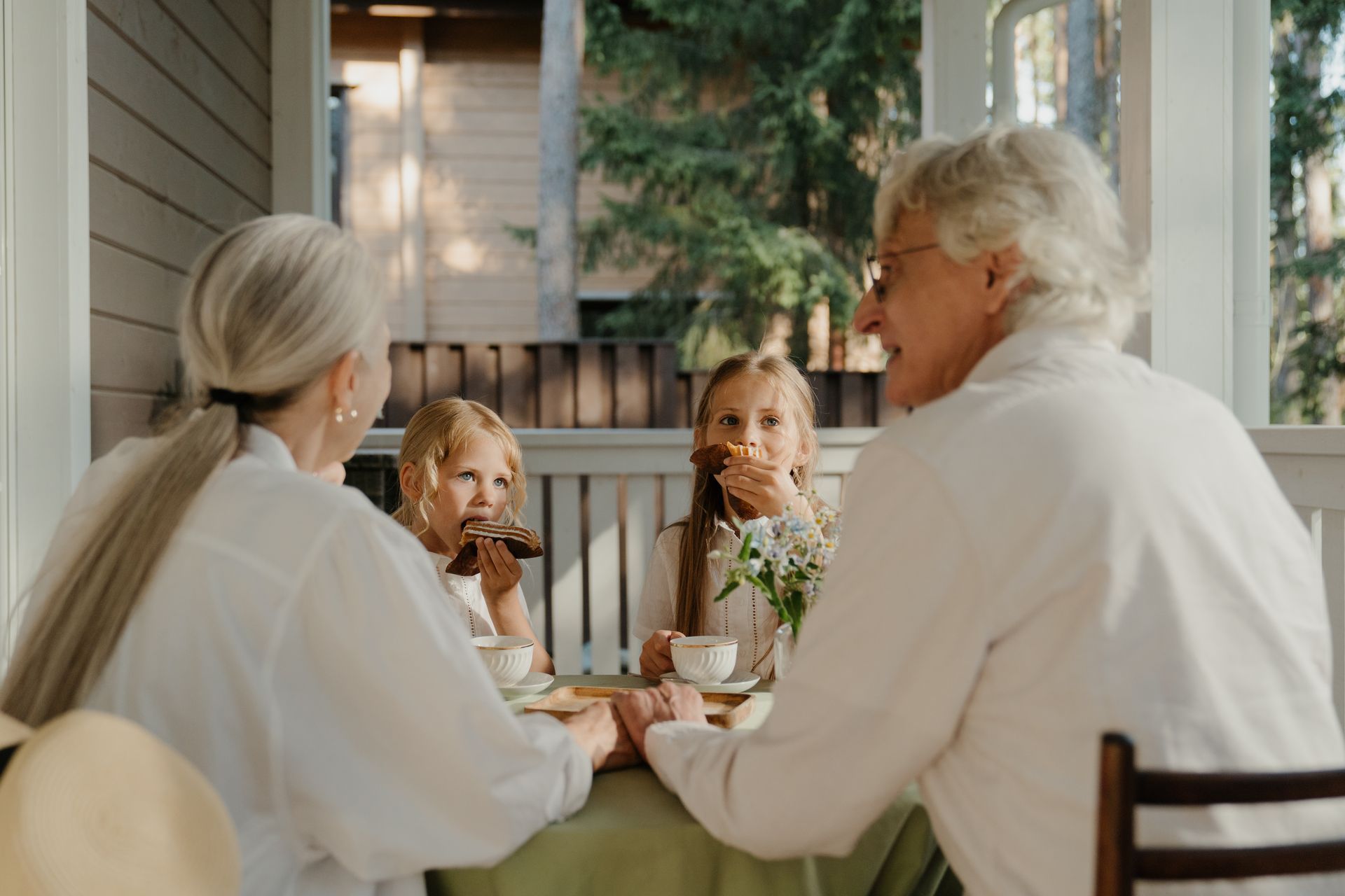 Three people sharing tea at a porch table, seen from behind, with flowers and a cozy outdoor setting