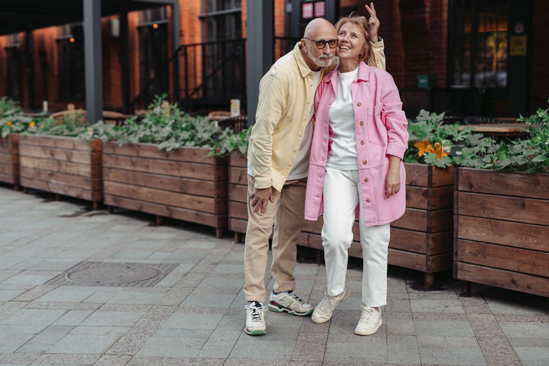Two people standing arm-in-arm on a city sidewalk beside wooden planters, one in pink and one in beige.