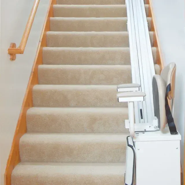 Stairlift installed near a window; beige seat, armrests, and safety belt.