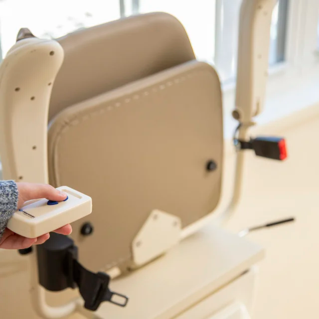Person operating a stairlift with a handheld remote. Beige seat with safety belt, window in background.