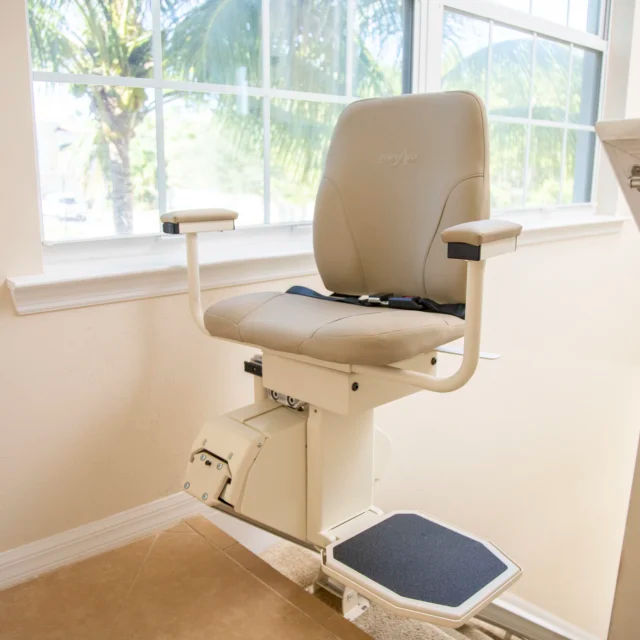 Stair lift with beige seat, armrests, and footrest, beside a window.
