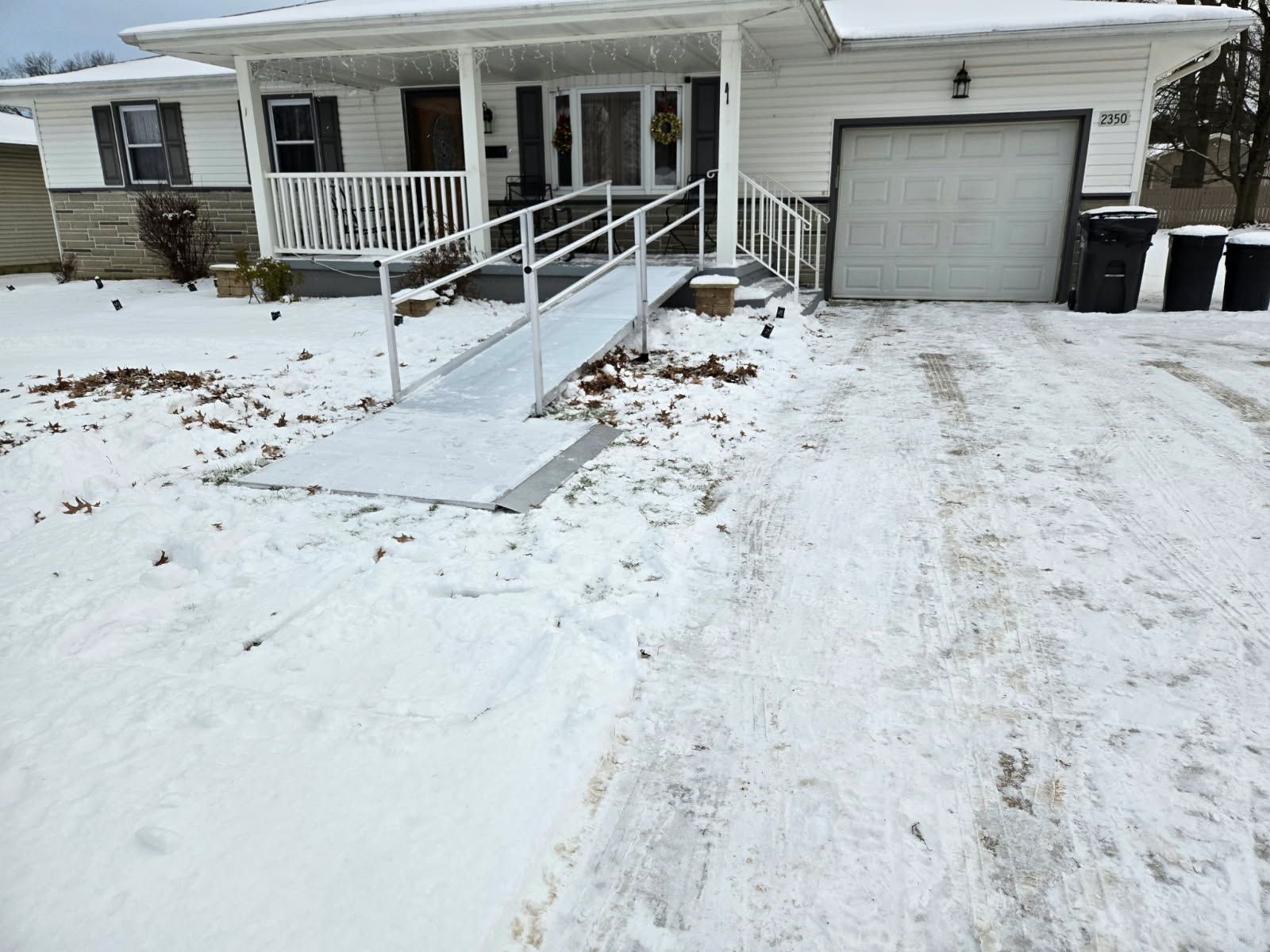 Snowy exterior of a white house with a metal wheelchair ramp leading to the front porch. The driveway is also snow-covered.