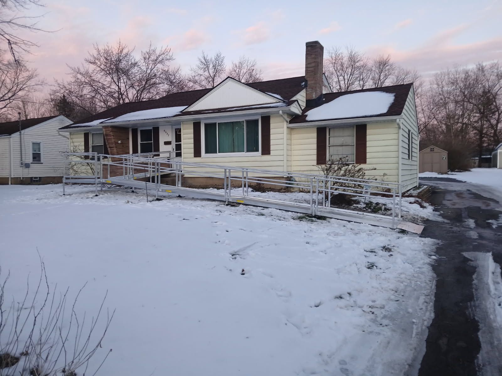 A one-story beige house with a snow-covered yard and a metal ramp leading to the entrance. The sky is pale pink.