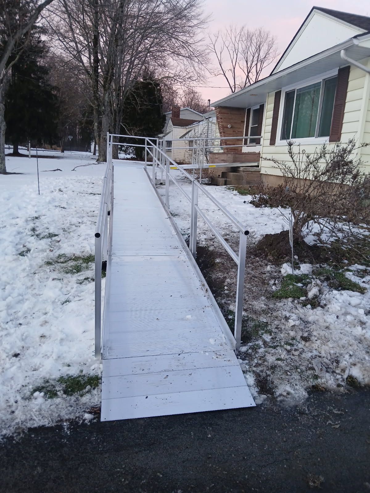 A long, aluminum ramp with handrails leads to the front door of a house covered in snow.