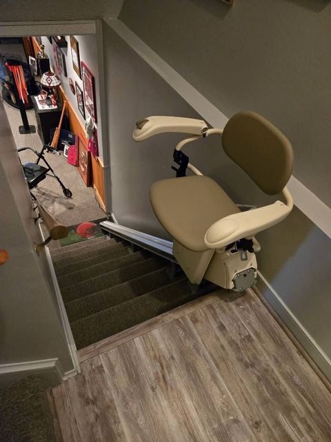 Stair lift installed on a staircase. Beige seat and armrests, gray walls and steps, wood-look flooring.