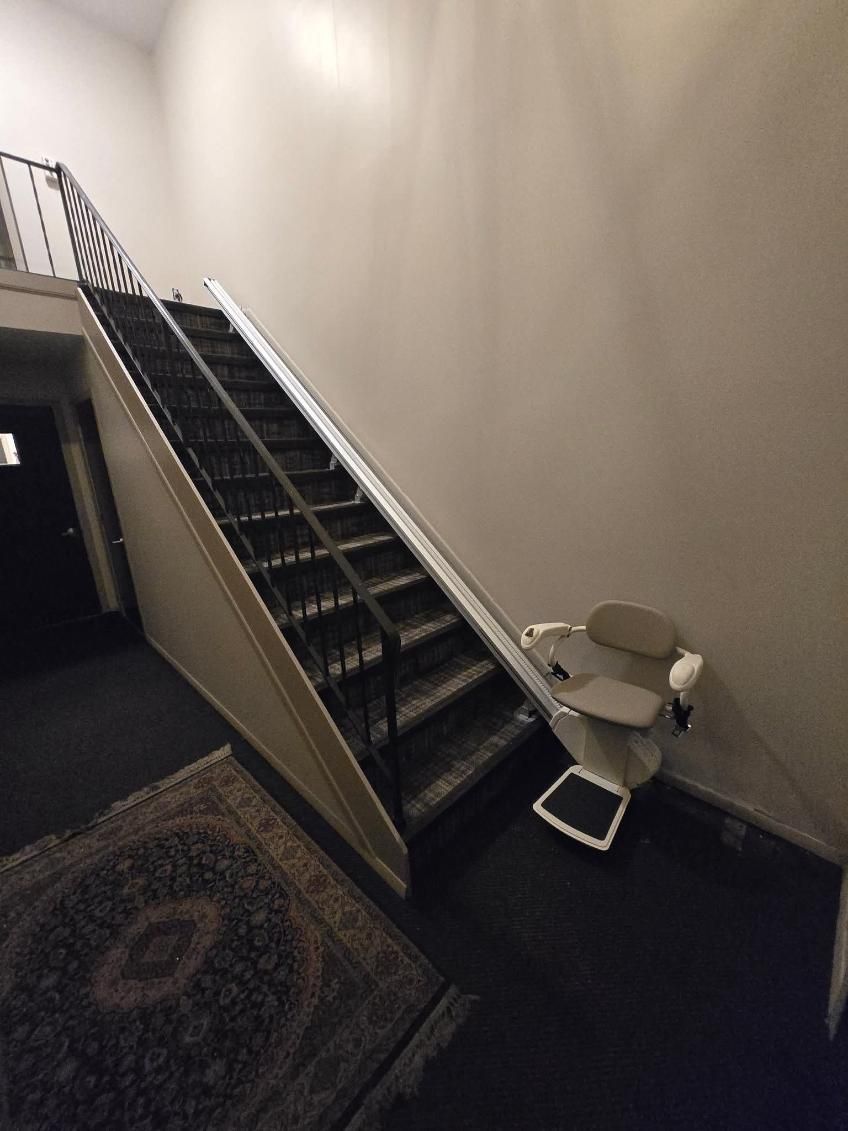 Staircase with a beige stairlift. Black stairs, white walls, and a decorative rug.