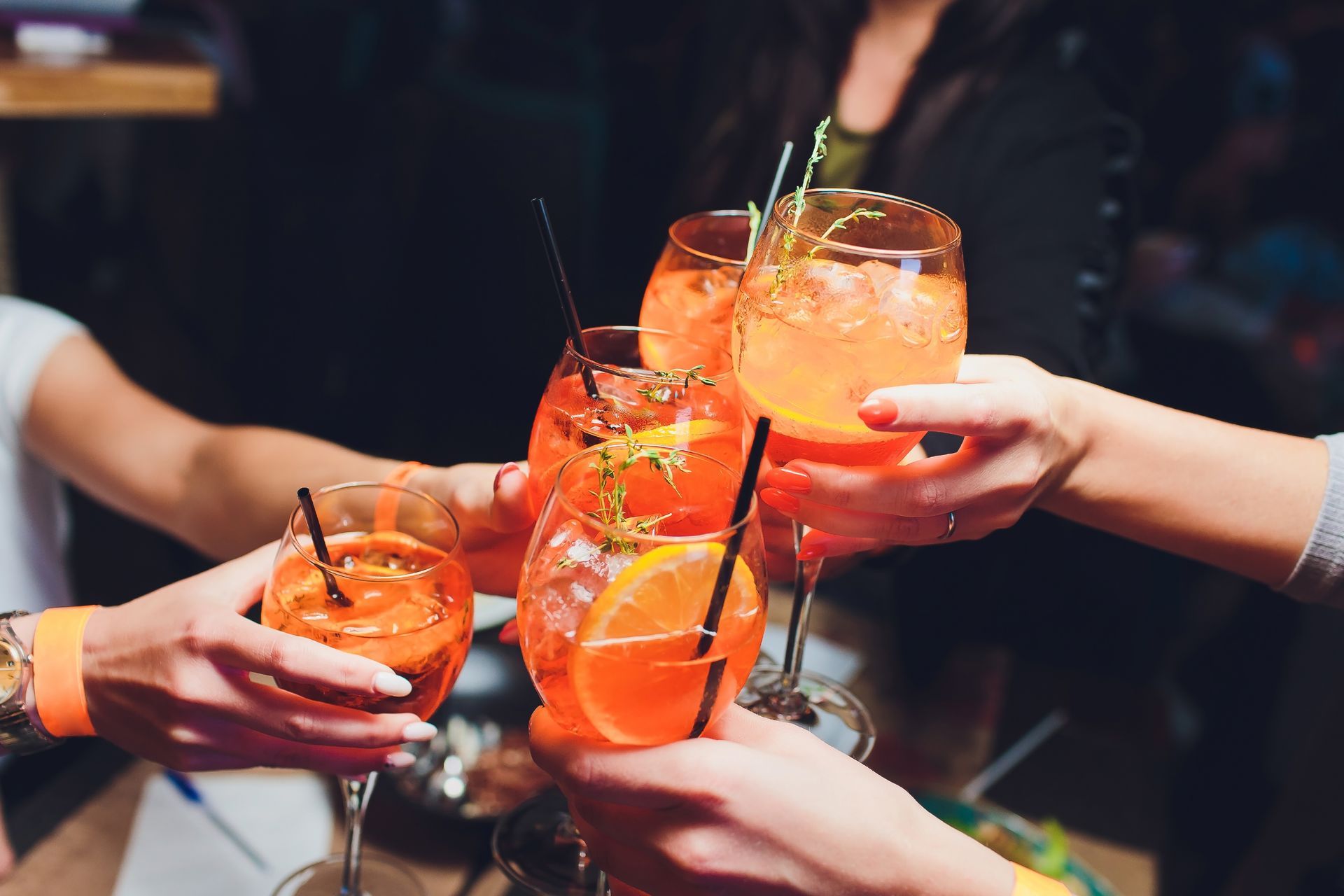 Hands Toasting Aperol Spritzes in a Dimly Lit Bar, Orange Drinks With Straws and Orange Slices — Chefs2Go in Mackay, QLD