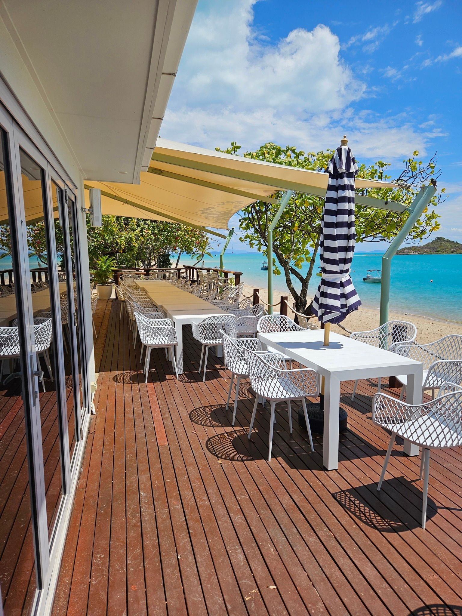 Outdoor Dining Area With Tables, Chairs, and Umbrella on a Wooden Deck Near a Beach — Chefs2Go in Woodwark, QLD