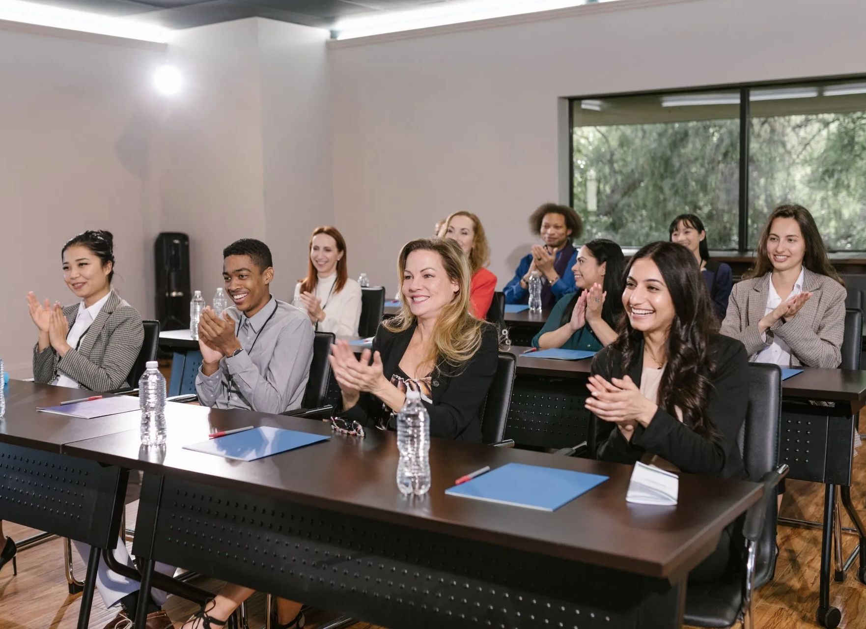 A group of people are sitting at tables in a classroom clapping.
