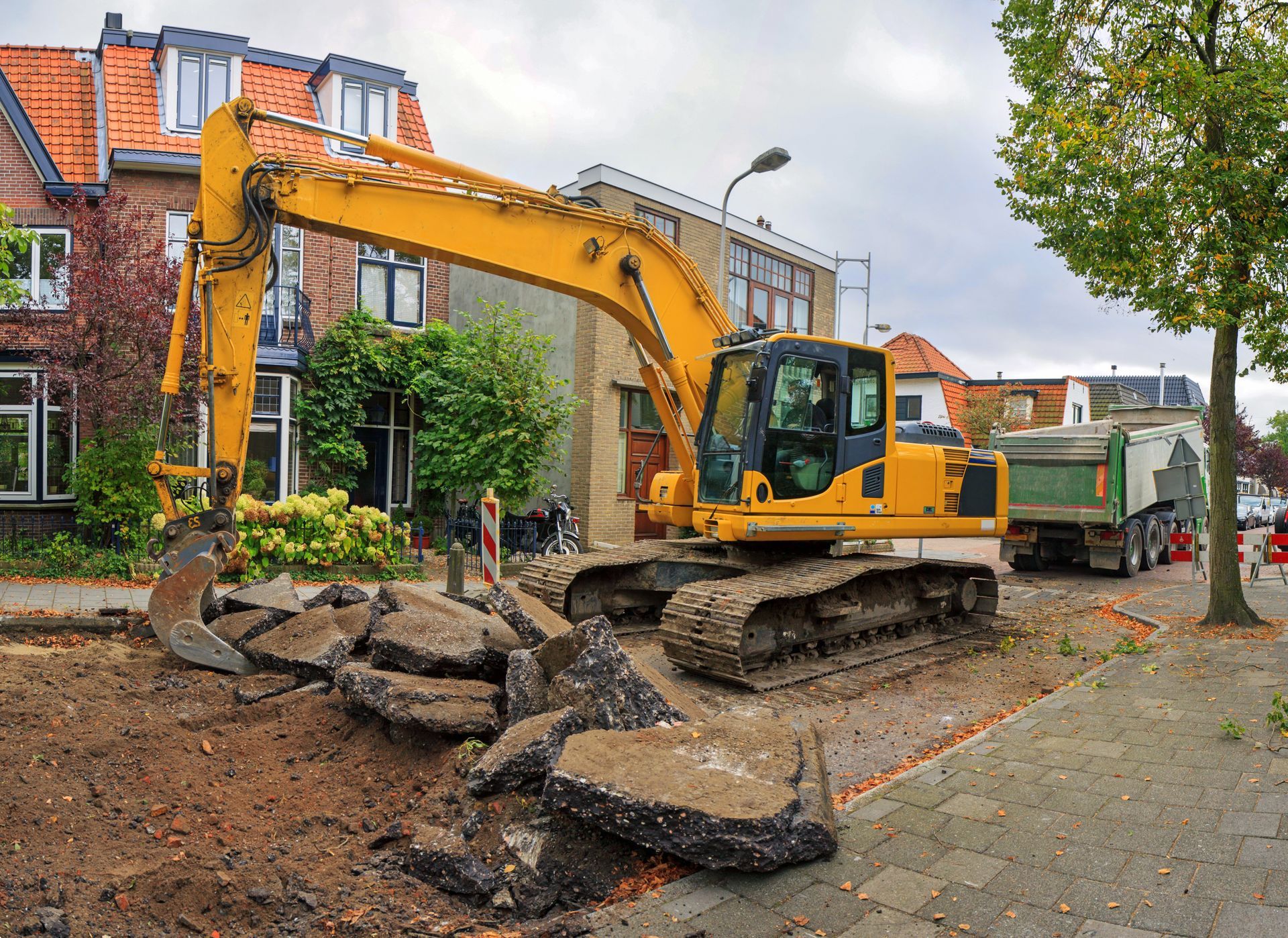 Yellow excavator tearing up asphalt on a residential street; a dump truck and houses are in the background.