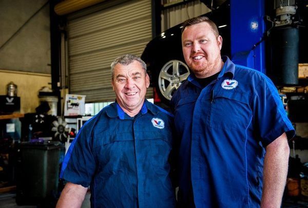 Two Men in Blue Mechanic Uniforms Smiling in A Garage — DK Mechanical Repairs in Warners Bay, NSW