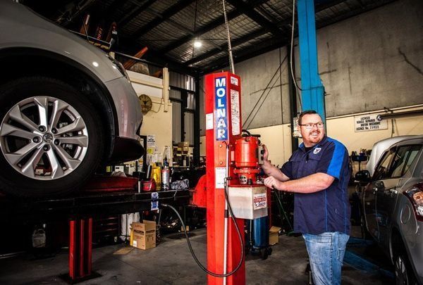 Mechanic in Auto Shop Smiling Operating Lift Under Car — DK Mechanical Repairs in Warners Bay, NSW