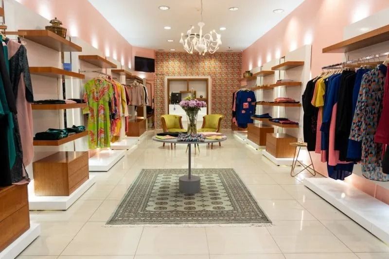 Tienda de ropa boutique luminosa con vestidos coloridos, estantes y una lámpara de araña bajo una suave iluminación rosa.