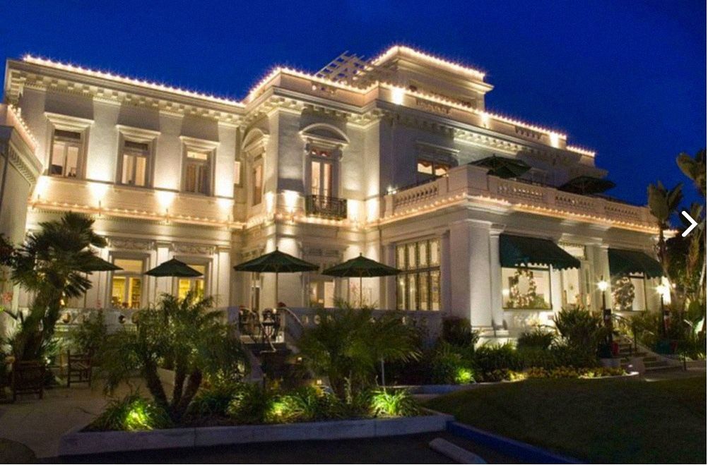 Elegant white mansion lit with string lights at night; outdoor dining area with umbrellas.