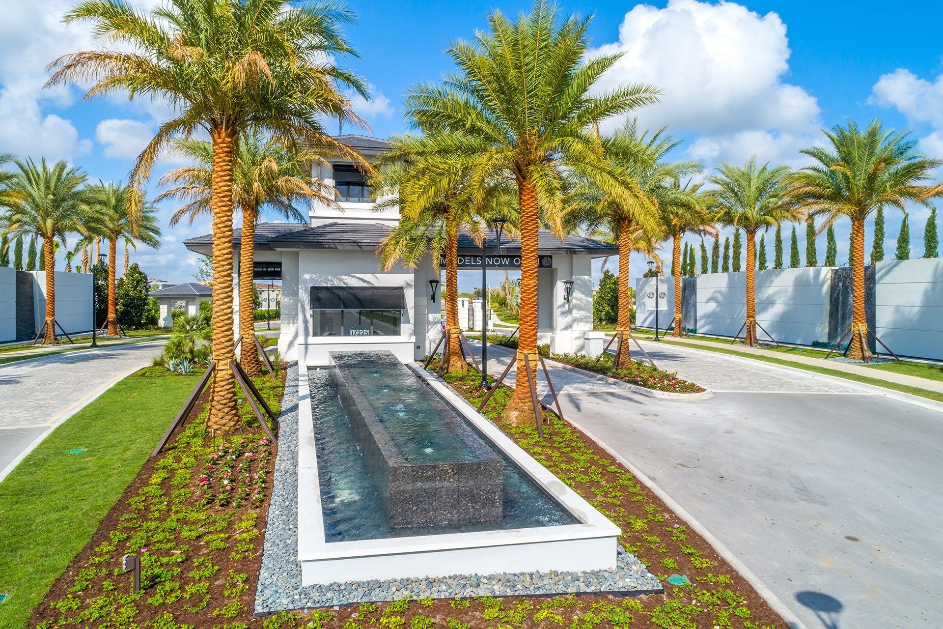 Gated entrance with fountain, palm trees, and modern building under a bright blue sky.