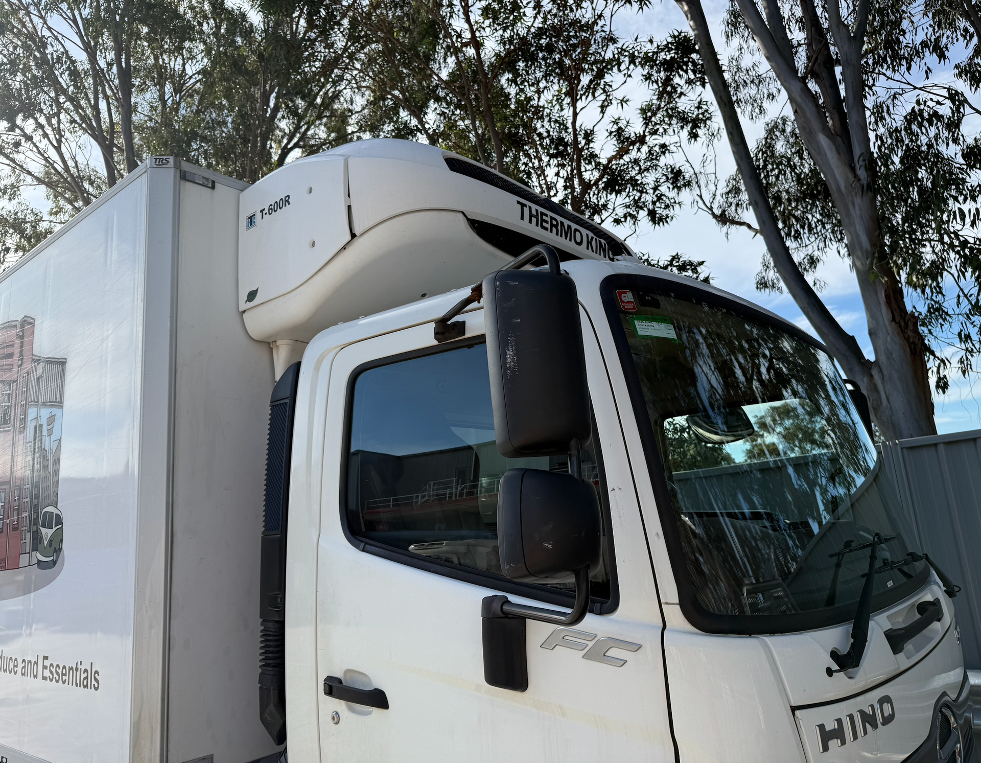 A white Hino refrigerated truck cab is parked outdoors under trees on a bright day.