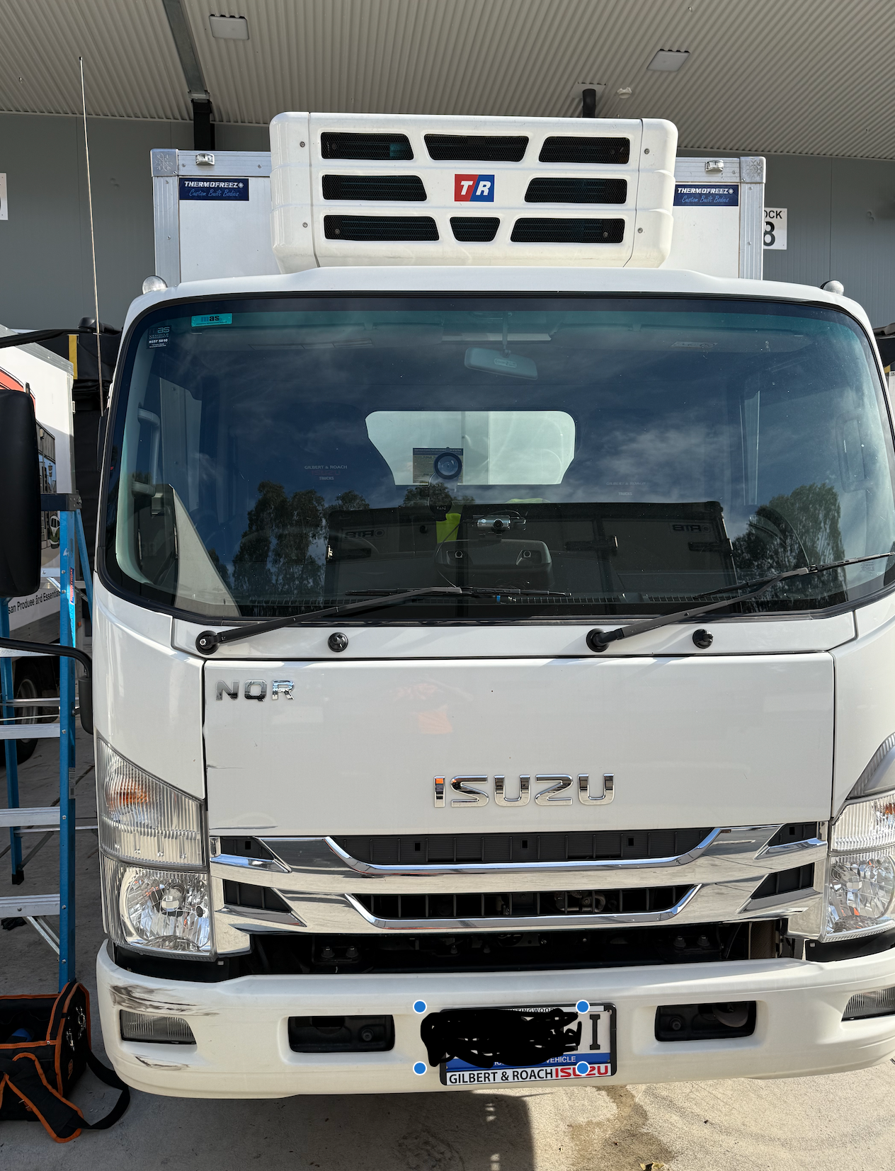 A white Isuzu NQR refrigerated box truck parked outdoors under a roof.