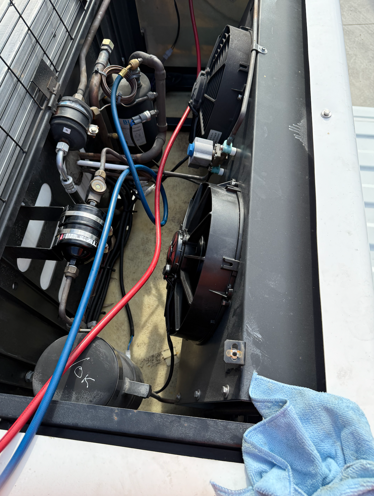 An open refrigeration unit showing internal components, including two black fans, copper piping, and red and blue hoses.