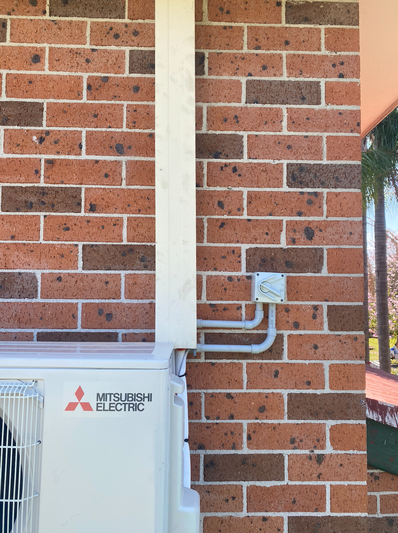 A Mitsubishi Electric air conditioner outdoor unit sits against a red brick wall next to white electrical conduits.