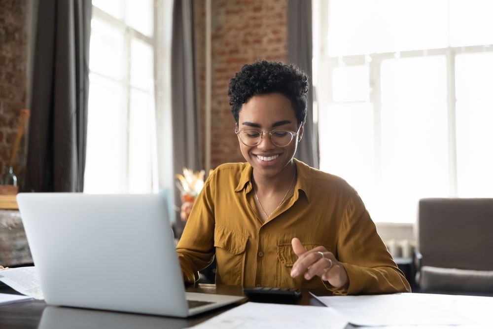 Person smiling at a laptop in a bright home office, with papers and a phone on the desk.