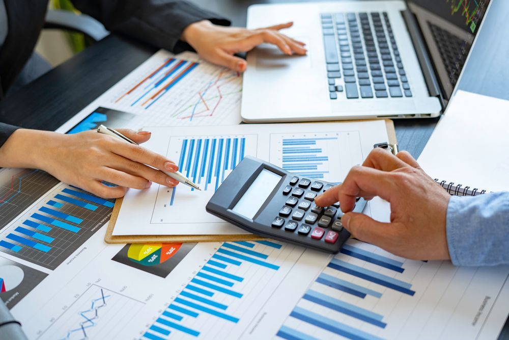 Hands using a calculator over business charts and reports on a laptop desk