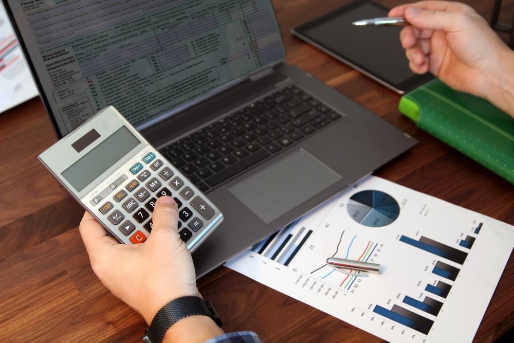 Hands using calculator and laptop beside printed charts on a desk