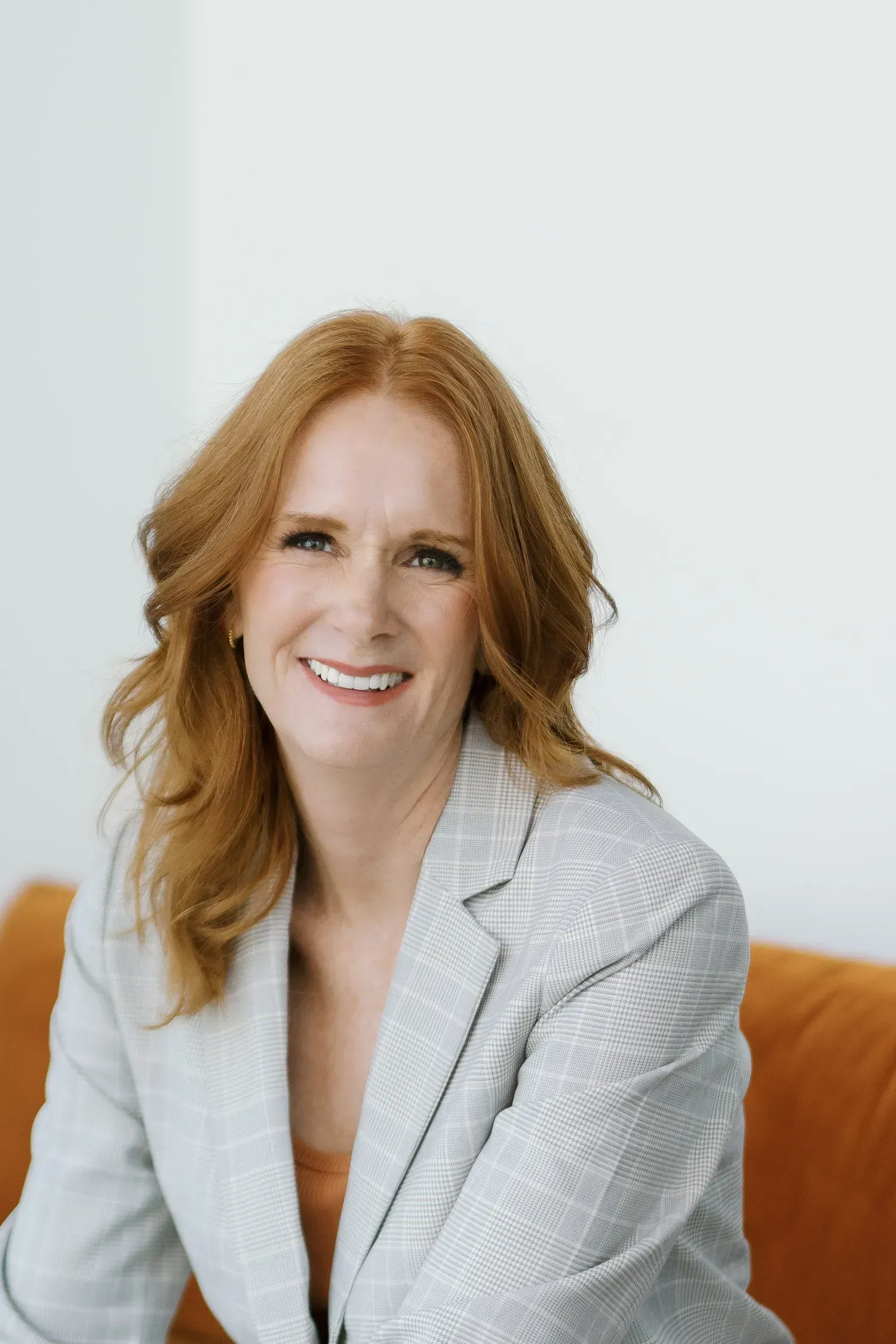 Smiling woman in a light gray blazer seated on an orange couch against a white background