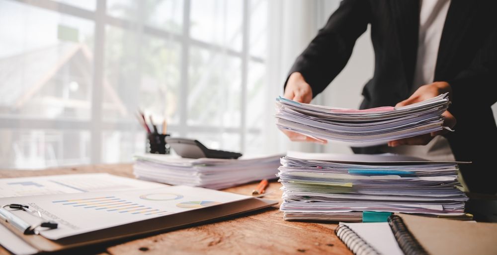 Person organizing a tall stack of documents on a wooden desk in an office workspace