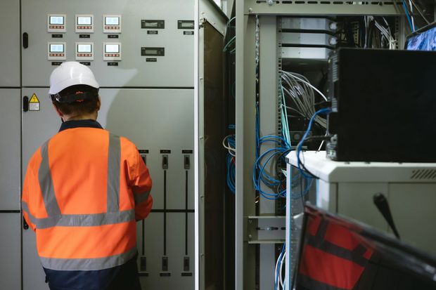 Tecnico con giubbotto di sicurezza arancione ispeziona il quadro elettrico e il cablaggio in un locale industriale.
