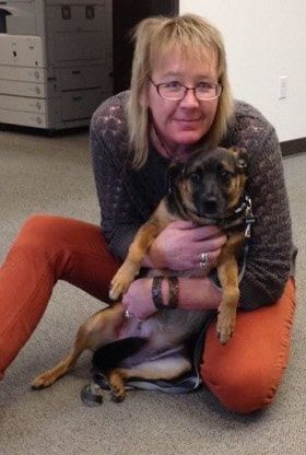 A person in an orange sweater and dark cardigan kneels on a carpeted floor, holding a small brown dog in their arms.