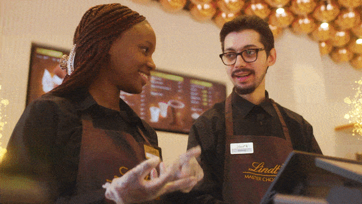 Two café staff members in black aprons and gloves smile while serving at a counter under warm lights.