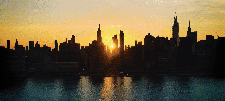 Silhouetted city skyline at sunset with a golden sun reflecting on the water