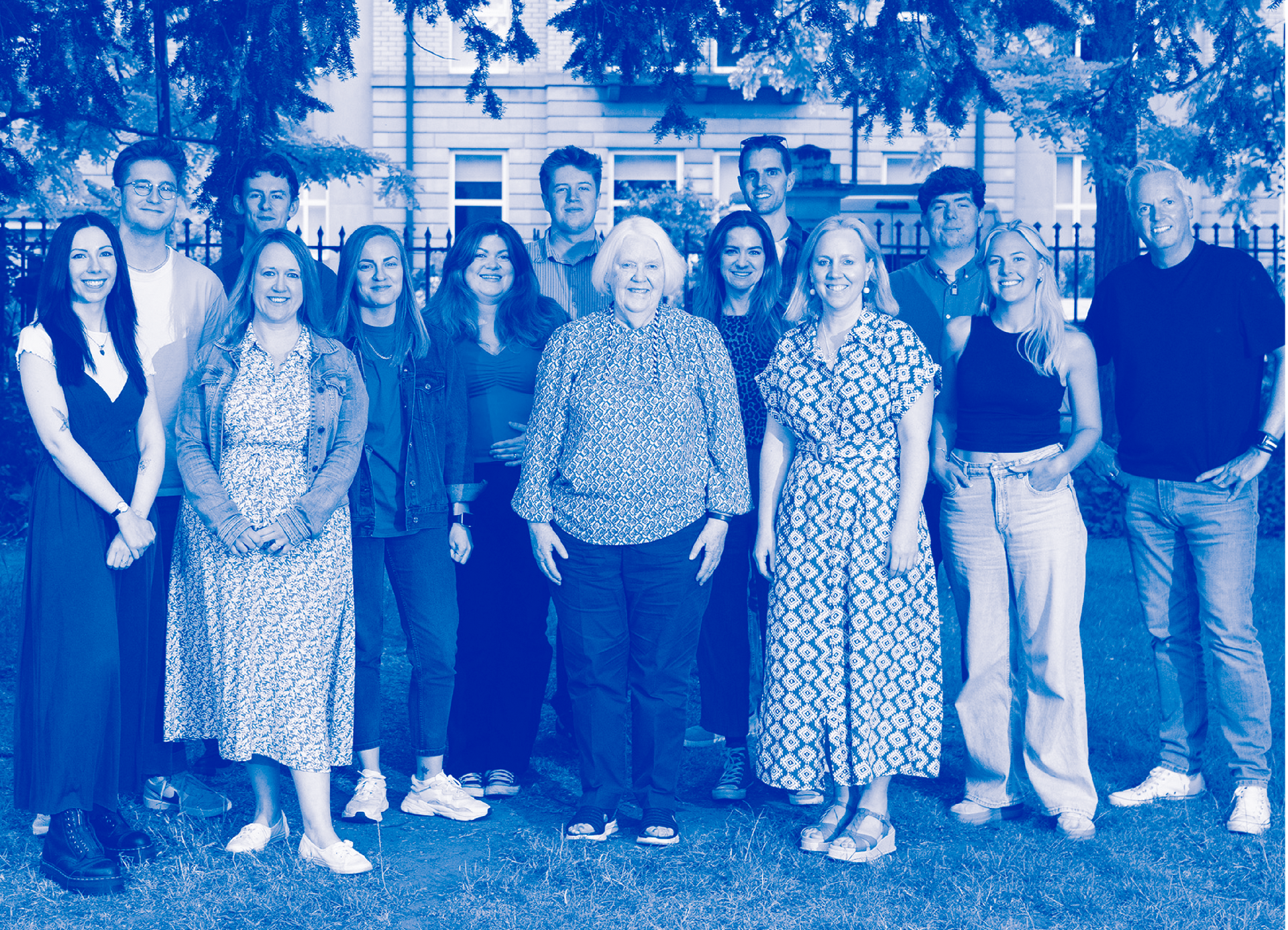 Group of people standing outdoors under trees in front of a building, tinted blue.