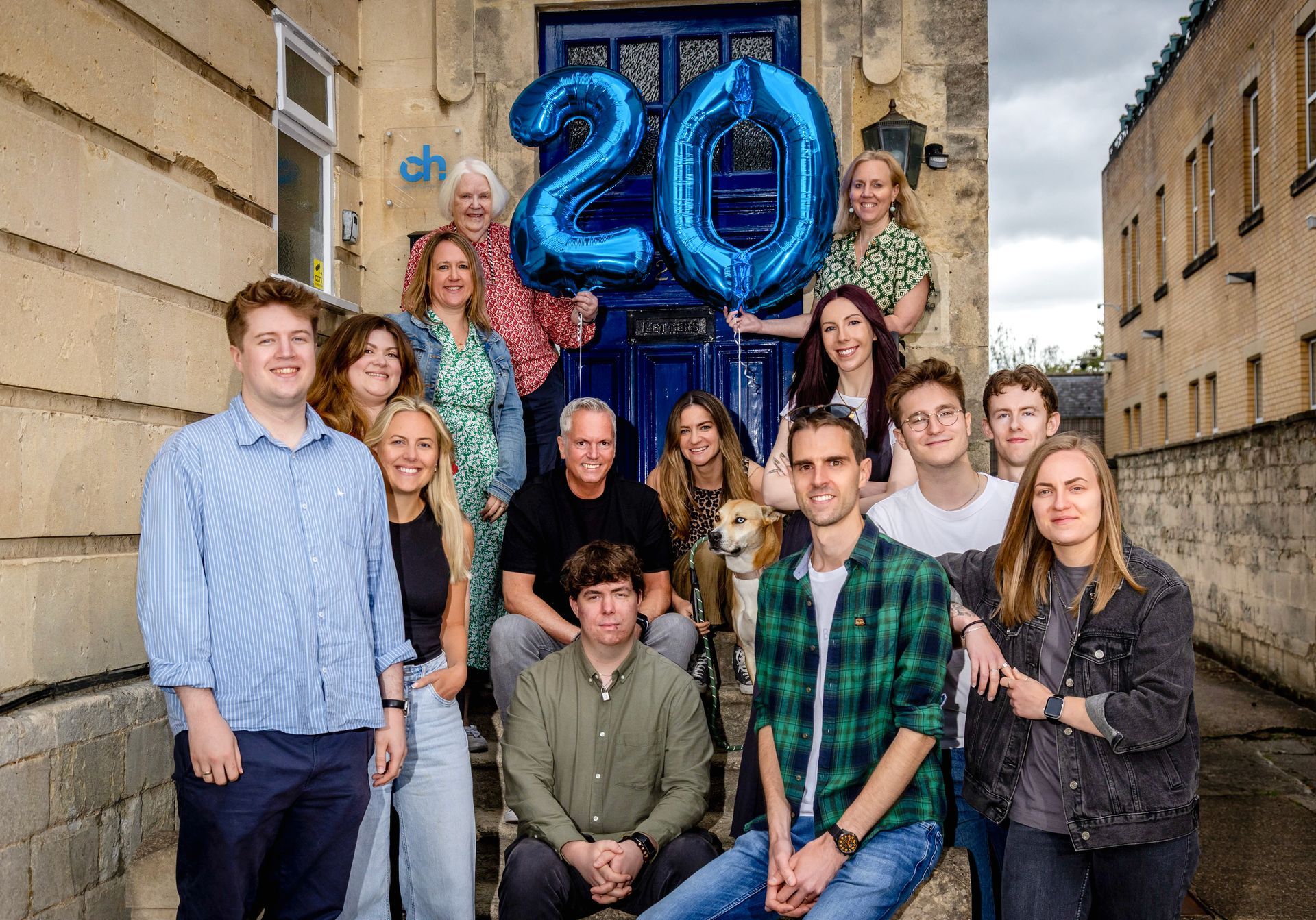 Group posing outside under blue balloons shaped “20” in a narrow street.