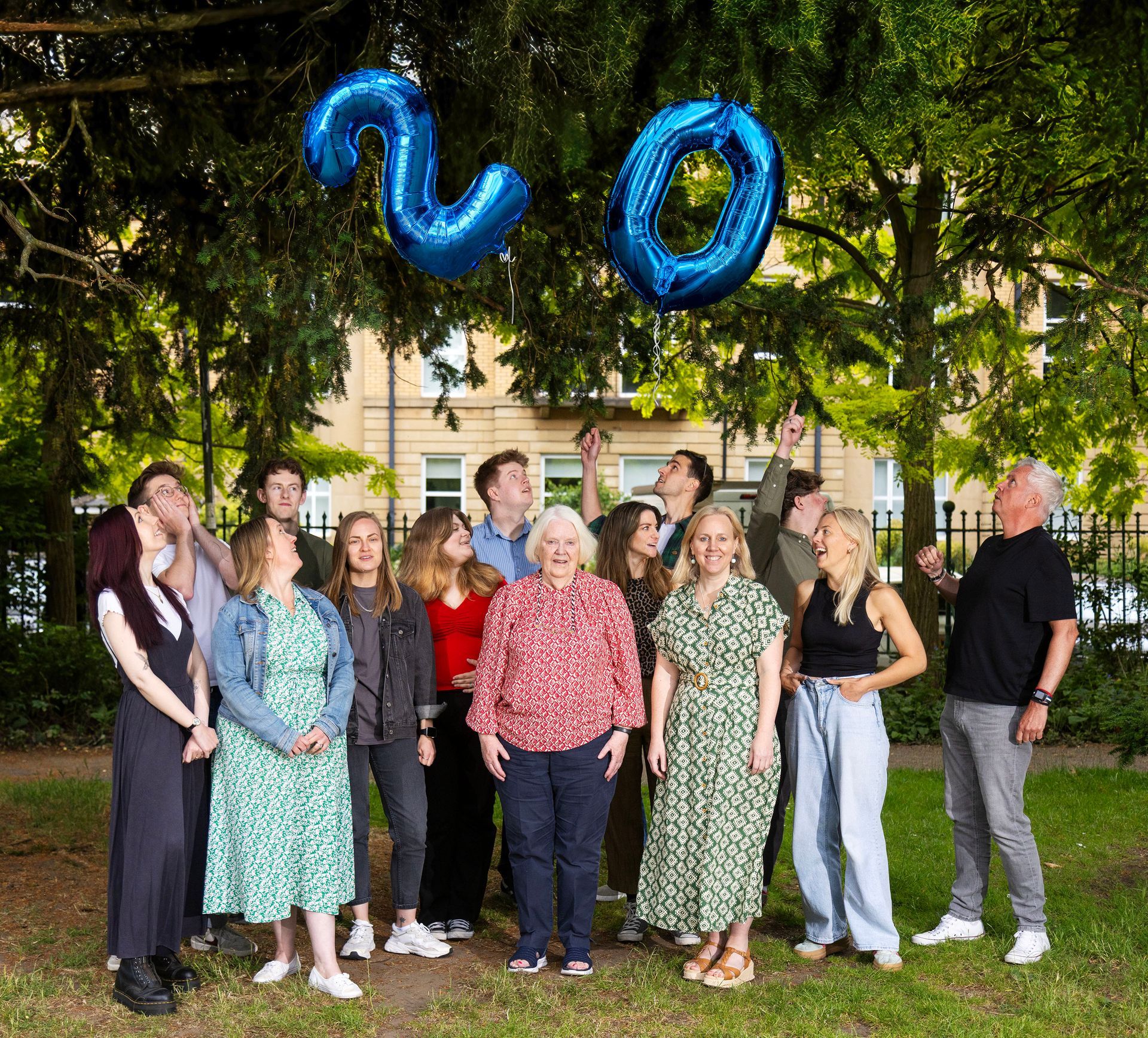 Group of people posing outdoors under blue “20” balloons in a park-like setting