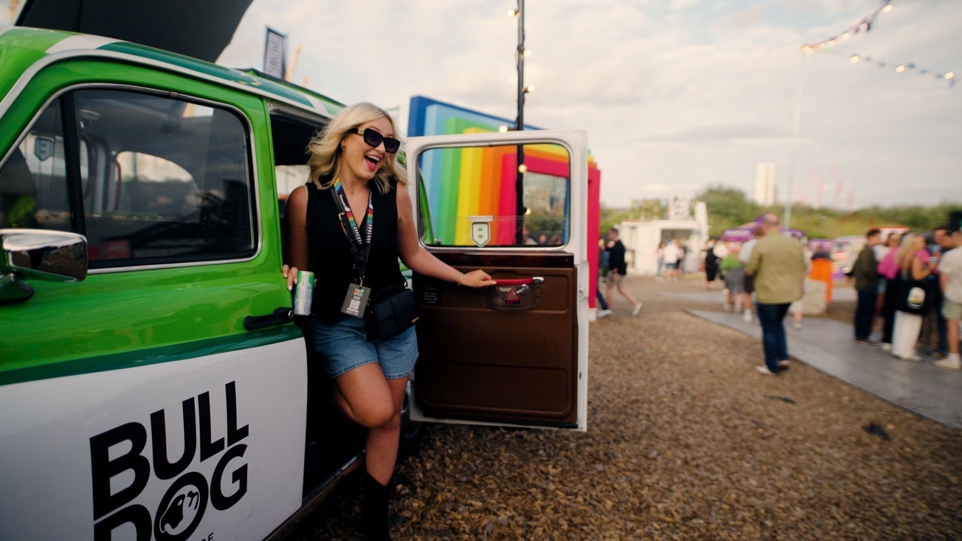 Woman stepping from a colorful taxi onto gravel, with a rainbow and people in the distance
