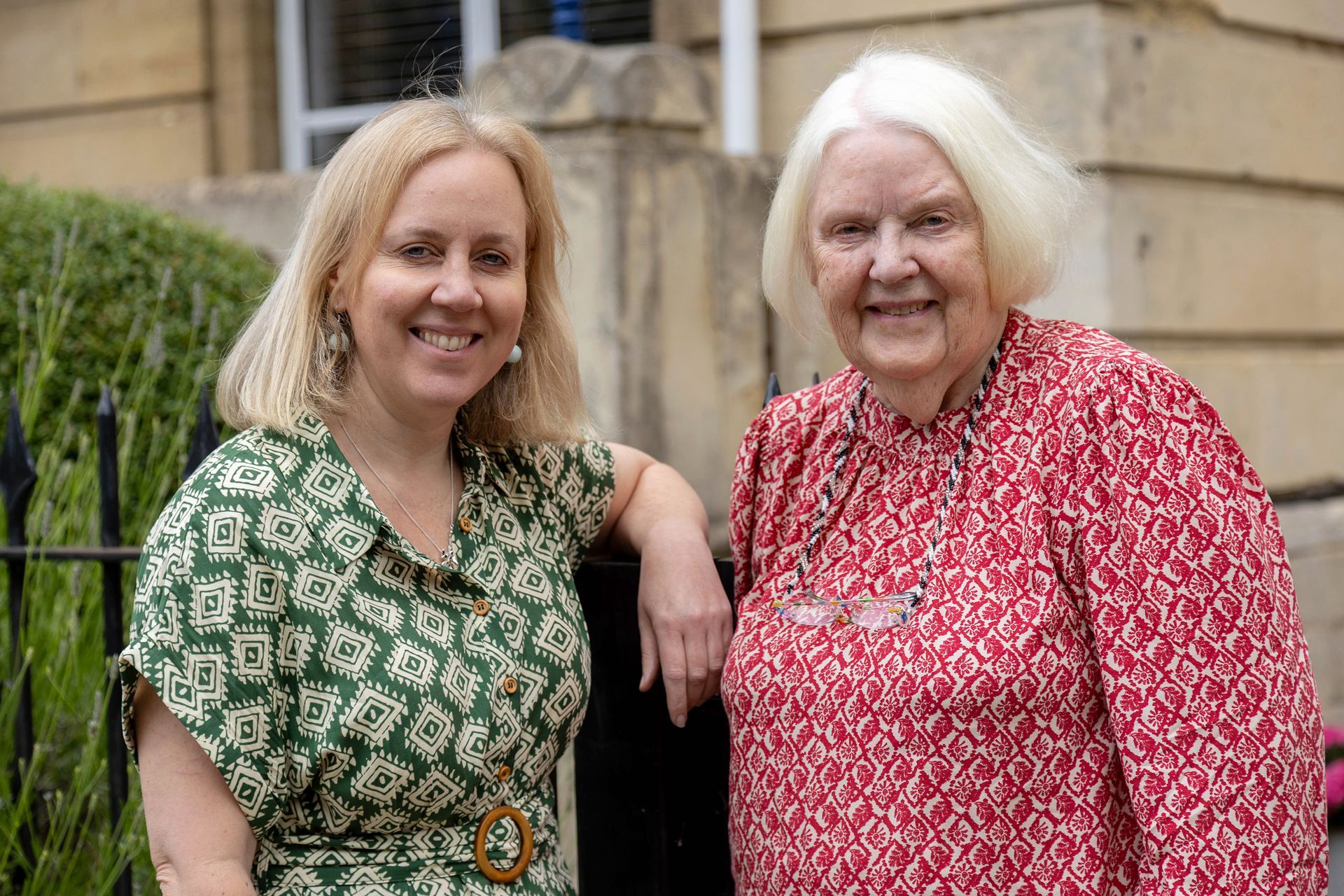 Two women standing outdoors, smiling beside a building, wearing green and red patterned tops.