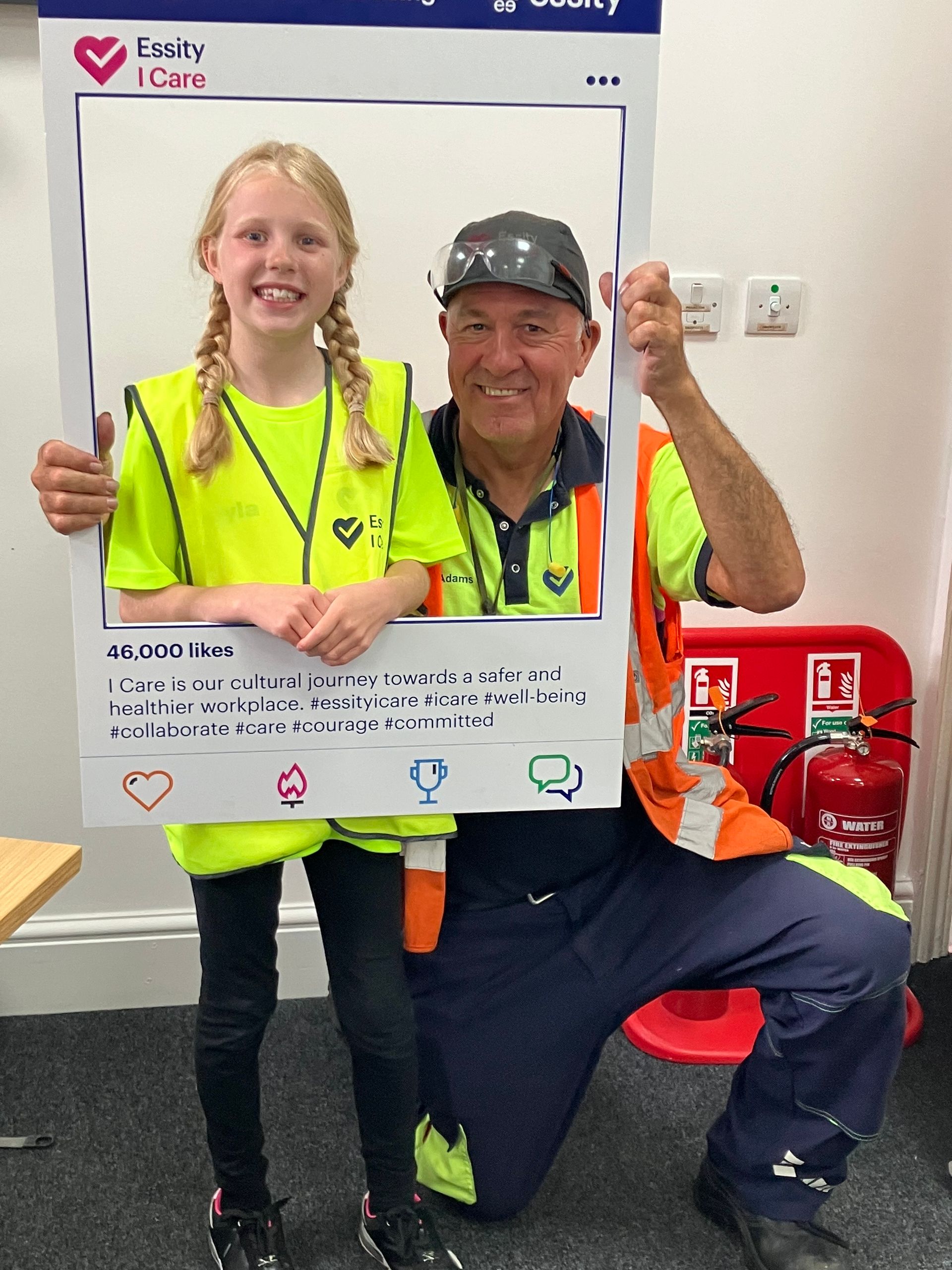 Two people in hi-vis vests smile while holding a large photo frame prop indoors.