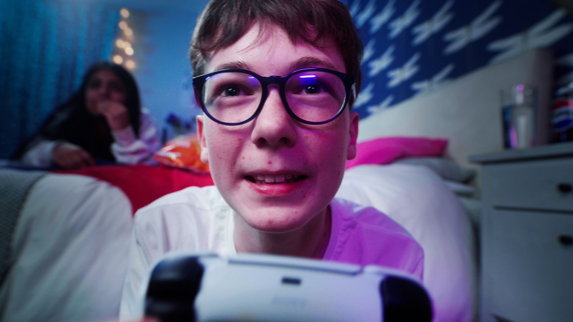 Close-up of a smiling person wearing glasses, holding a games console controller in a bedroom with someone blurred in the background.